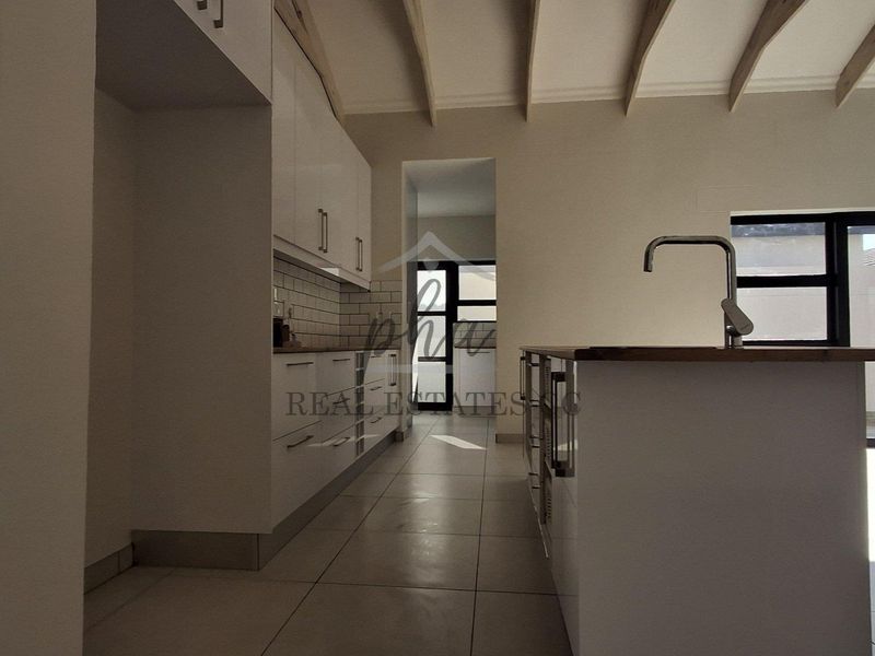 Modern kitchen featuring white cabinetry, a central island with a sink, subway tile backsplash, and exposed ceiling beams, leading to an outdoor area.