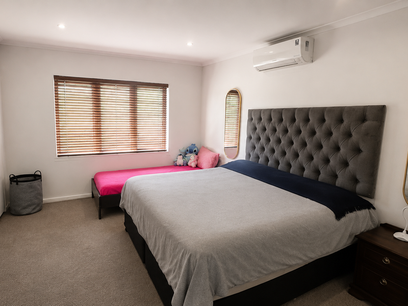 A well-maintained bedroom featuring neutral carpeting, built-in wardrobes, a split-system air conditioner, and large windows with venetian blinds.