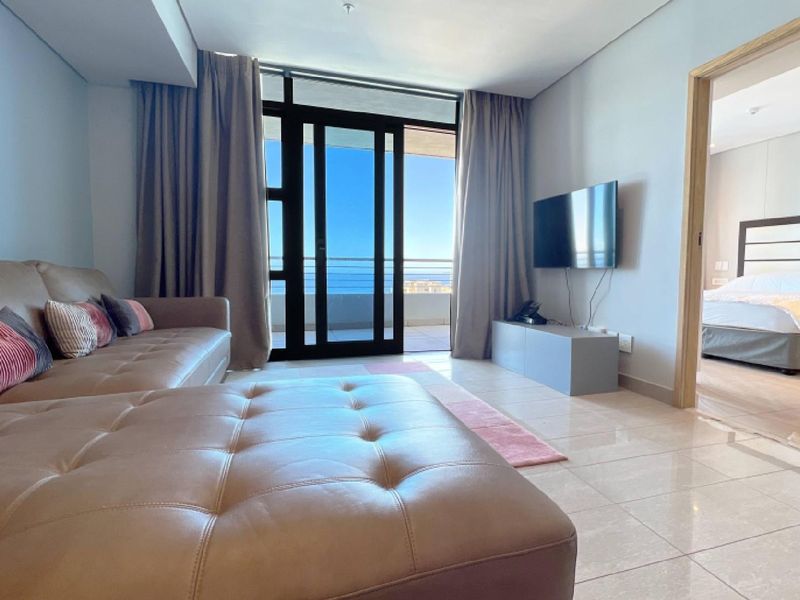 A bright living room featuring tiled floors, a recessed ceiling, and a large sliding glass door leading to a balcony with ocean views.