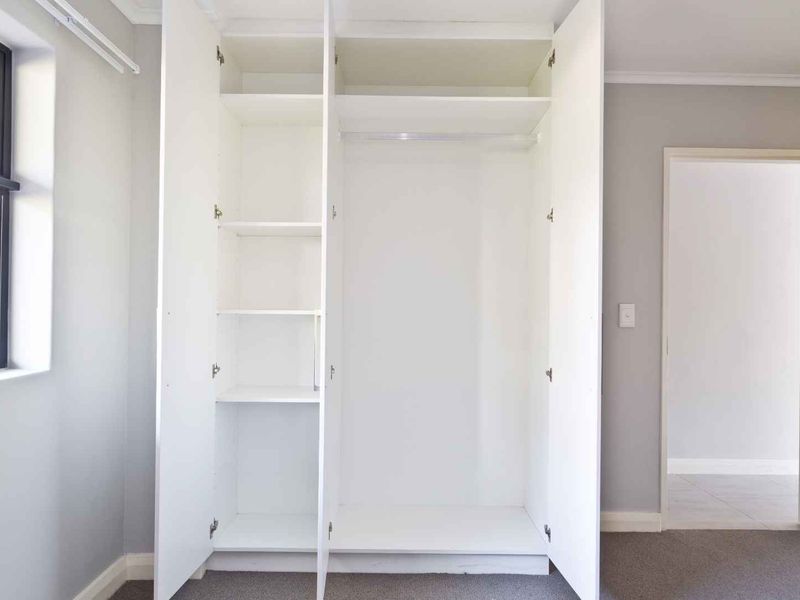 A bright, empty bedroom featuring a large white built-in wardrobe with shelving and hanging space, neutral grey walls, and carpeted flooring.