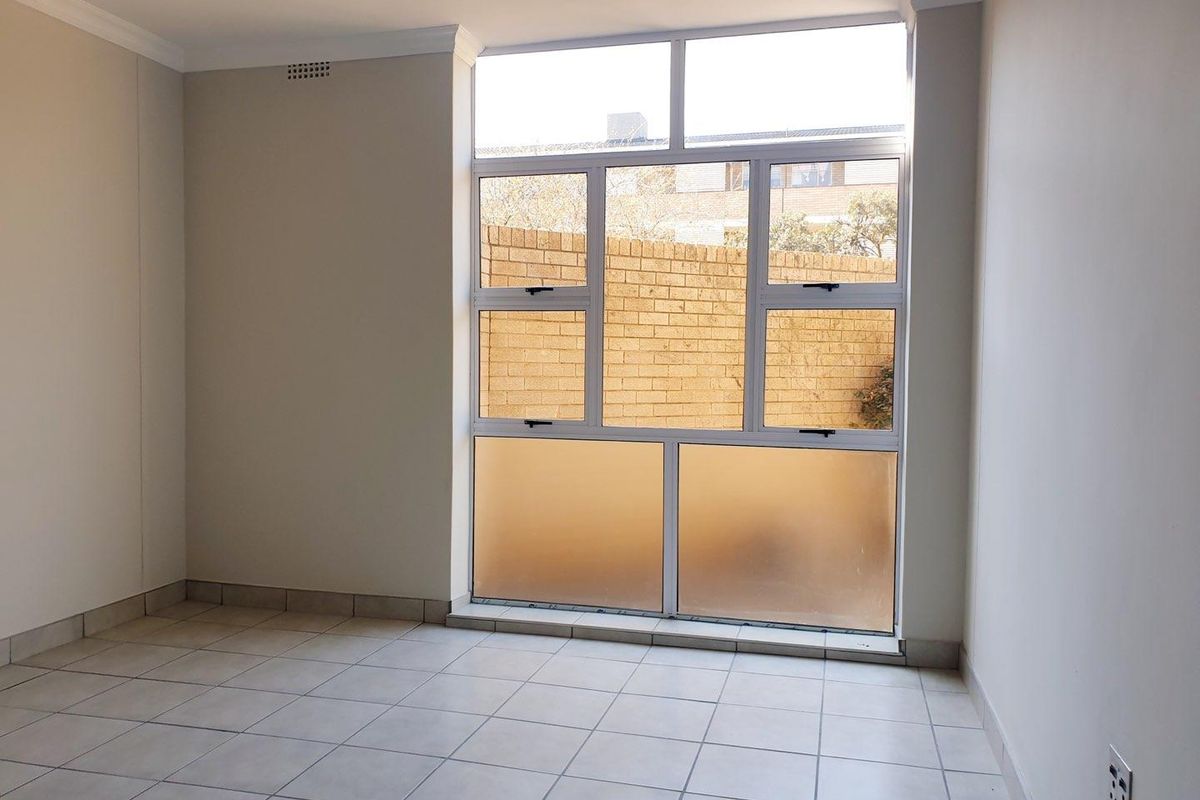 A bright, empty bedroom featuring neutral tiled flooring, clean walls, and large aluminium-framed windows providing natural light.