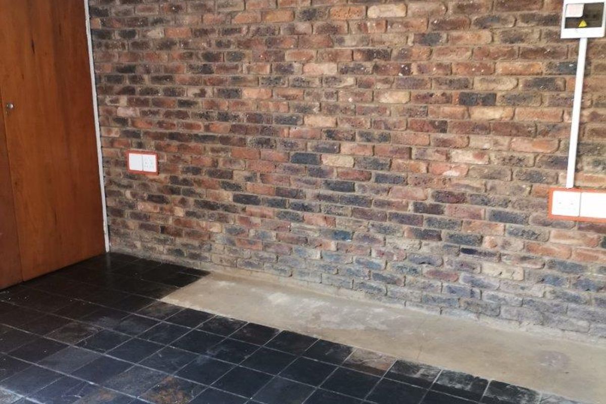 A garage space featuring an exposed brick wall, slate tile flooring, and a built-in wooden cupboard, showing signs of wear and dated finishes.