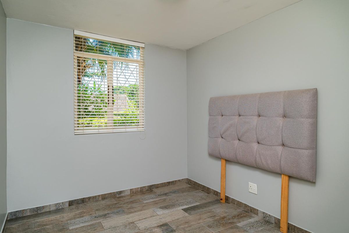 A bright, empty bedroom featuring modern wood-look tiled flooring, neutral grey walls, and a window fitted with venetian blinds.