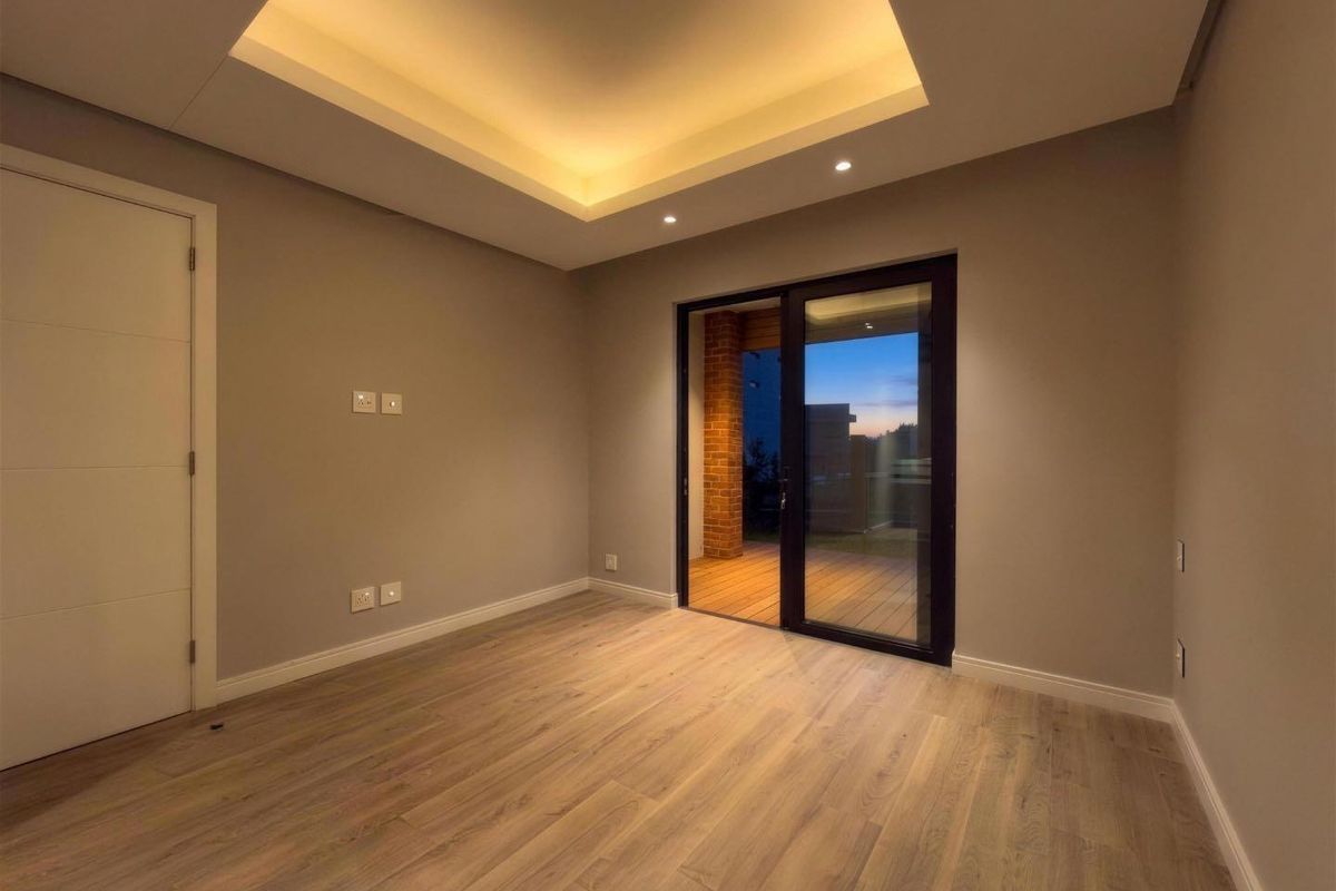 A modern, empty bedroom featuring a recessed ceiling with cove lighting, laminate flooring, and a sliding glass door leading to an outdoor area.