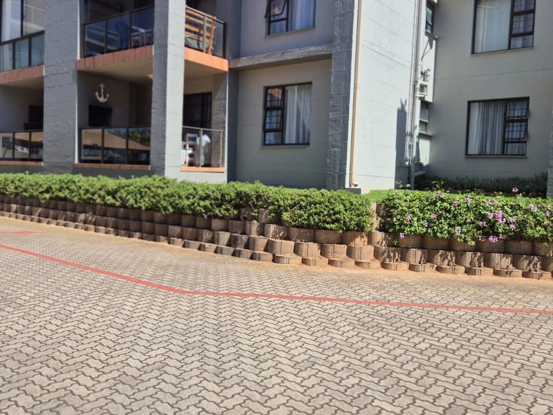 Exterior view of an apartment complex featuring a paved driveway, a tiered retaining wall with manicured hedges, and private balconies.