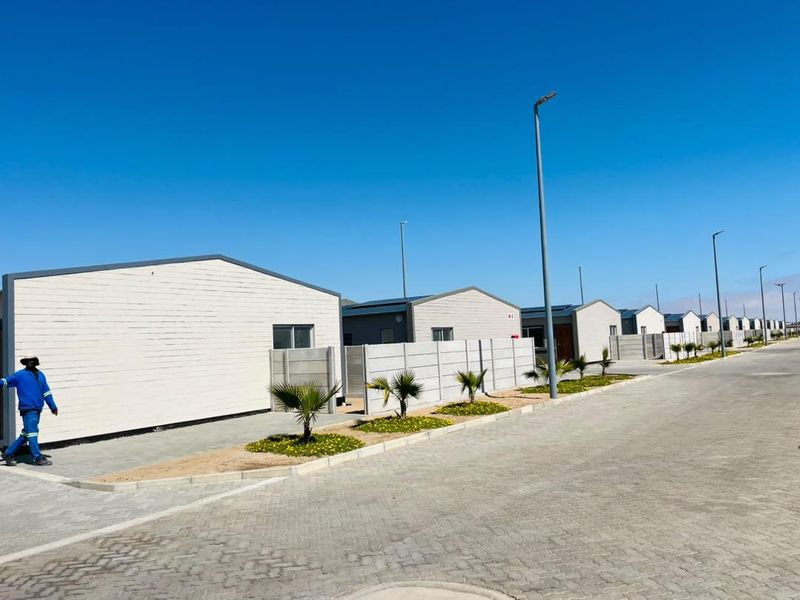 A street-level view of a modern townhouse development featuring uniform architectural design, paved driveways, and established boundary walls under a clear blue sky.