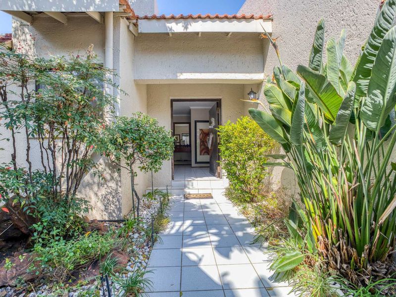 A welcoming front entrance featuring a tiled walkway leading to the main door, framed by established garden landscaping and stucco exterior walls.
