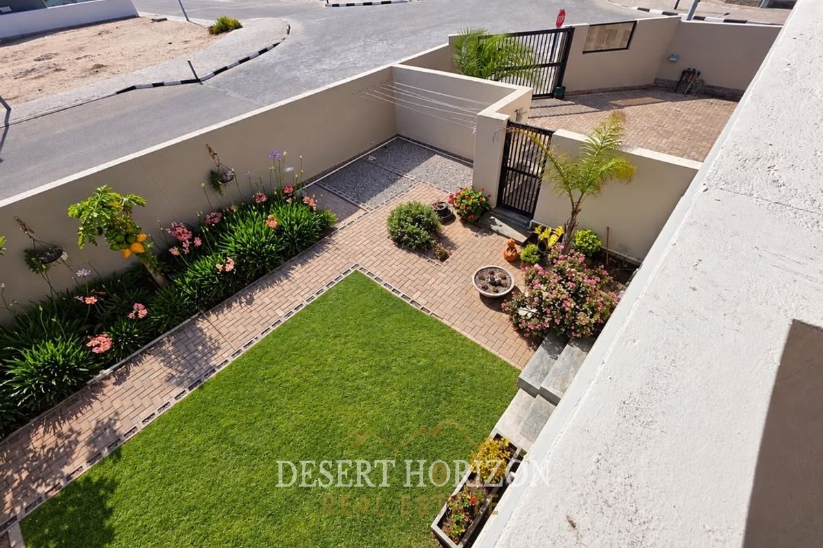 An elevated view of a well-maintained rear garden featuring a manicured lawn, paved pathways, boundary walls, and a private gated driveway entrance.