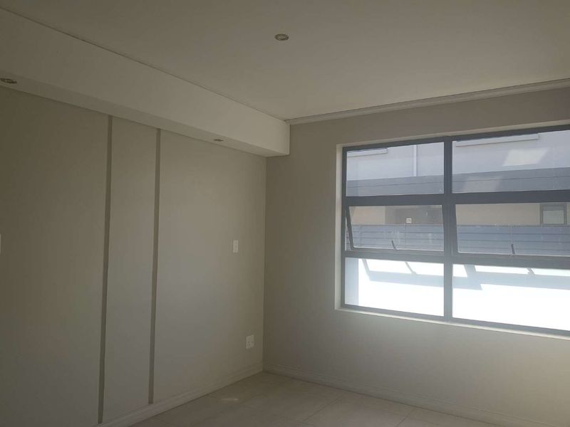 A bright, empty bedroom featuring neutral wall tones, tiled flooring, and modern aluminium window frames with recessed ceiling lighting.