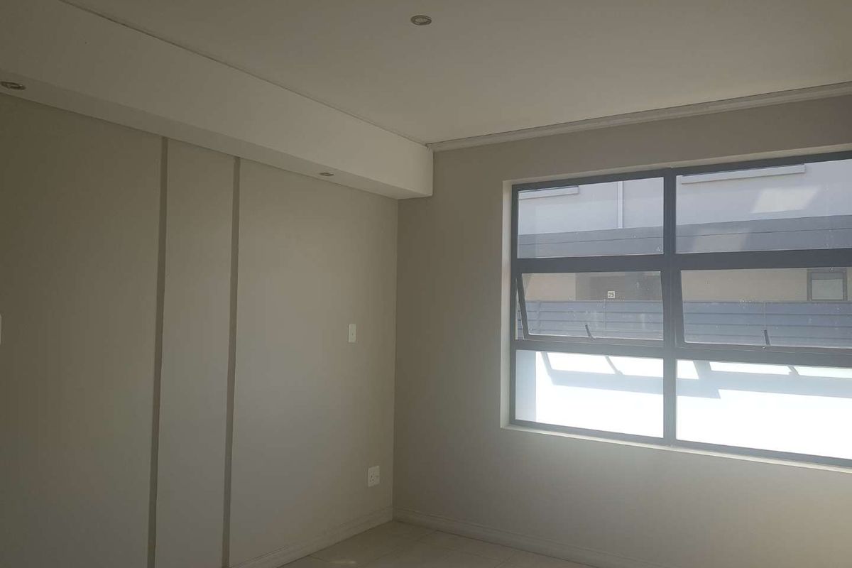 A bright, empty bedroom featuring neutral wall tones, tiled flooring, and modern aluminium window frames with recessed ceiling lighting.