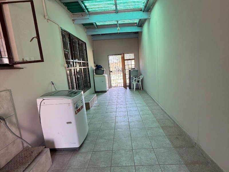 A functional laundry area featuring tiled flooring, exposed rafters with translucent roof sheeting, and direct access to the exterior via a security gate.