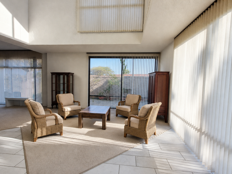 Spacious living area featuring double-height ceilings, large windows with vertical blinds, and a combination of tiled and carpeted flooring.