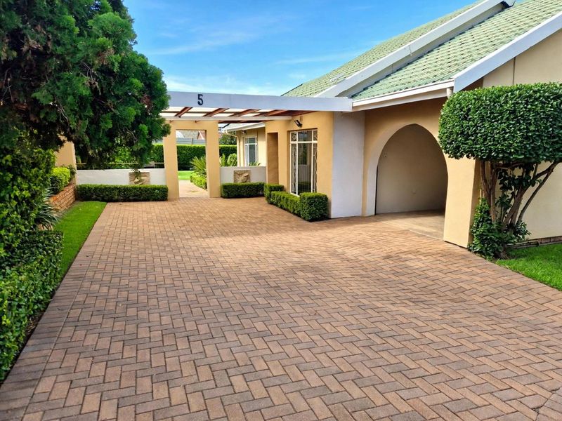 A well-maintained front exterior featuring a paved driveway, a covered pergola entrance, and an arched architectural detail with manicured garden hedges.