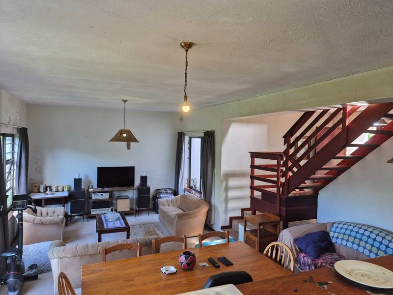 Open-plan living and dining area featuring a prominent wooden staircase, pendant lighting, and large windows, offering a spacious layout with potential for modernization.