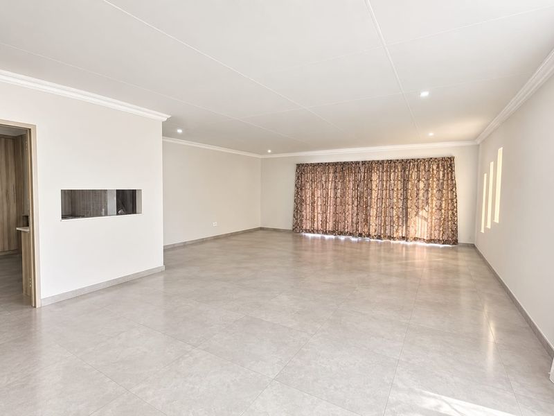 A spacious, empty living area featuring neutral tiled flooring, recessed ceiling lighting, and a convenient pass-through window connecting to the adjacent room.