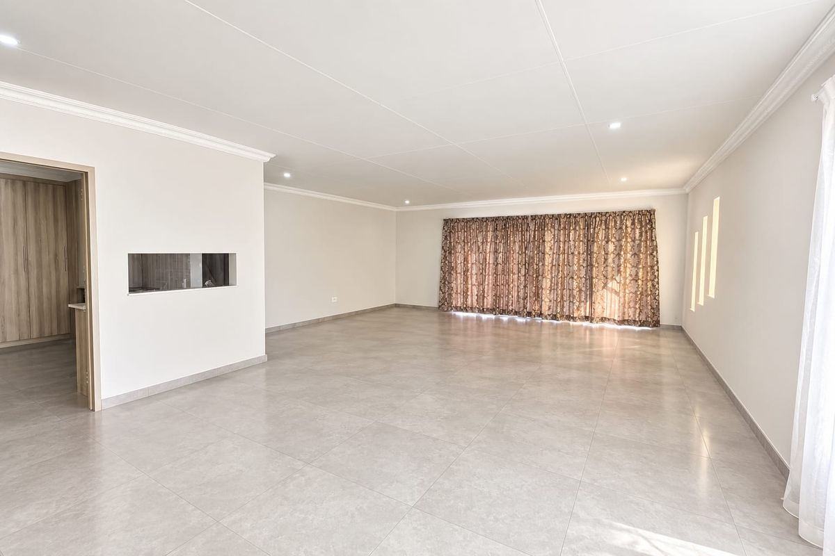 A spacious, empty living area featuring neutral tiled flooring, recessed ceiling lighting, and a convenient pass-through window connecting to the adjacent room.