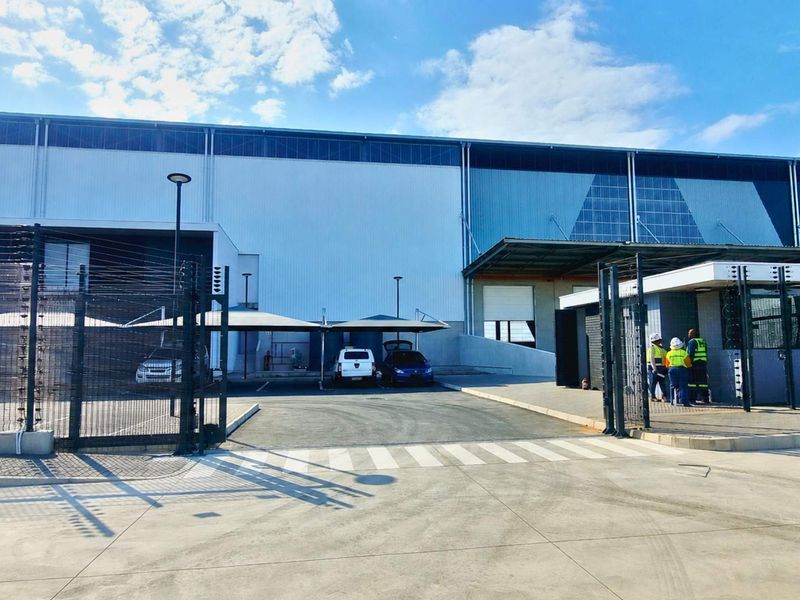 Front view of an industrial facility featuring a secure gated entrance, guard house, covered parking bays, and large-scale metal cladding exterior.