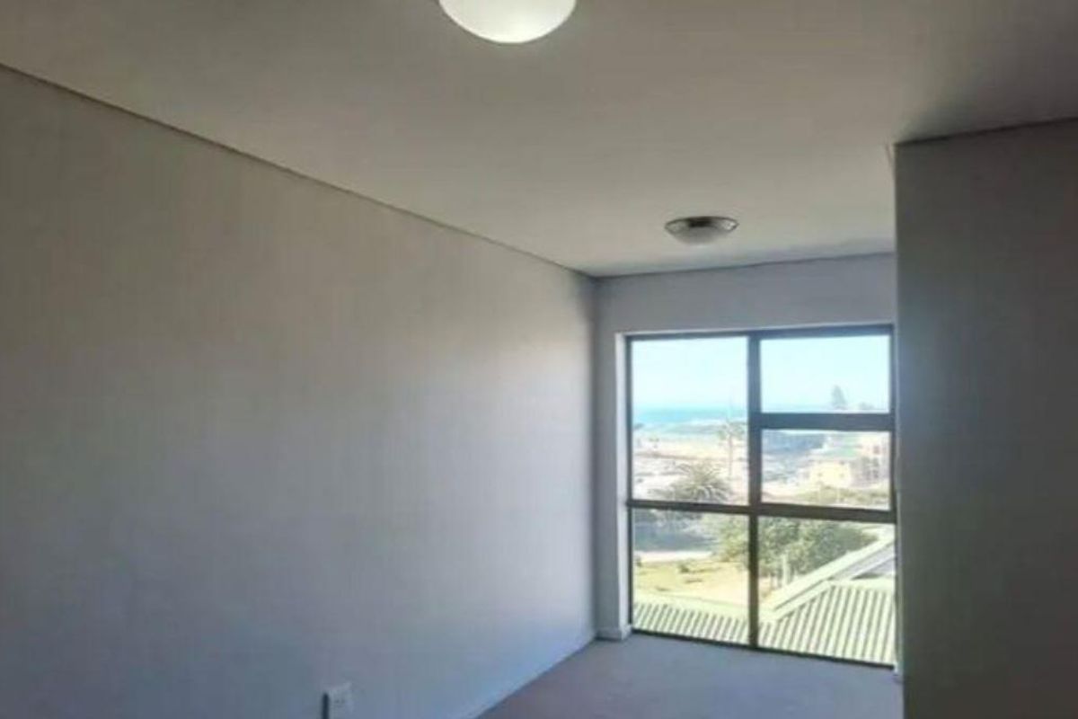 A bright, empty bedroom featuring neutral walls, carpeted flooring, and a large window offering a scenic coastal view.