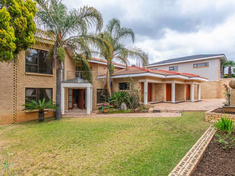 Spacious rear exterior view featuring a face brick facade, covered patio area, upper balcony, and a well-maintained garden with a stone fountain feature.