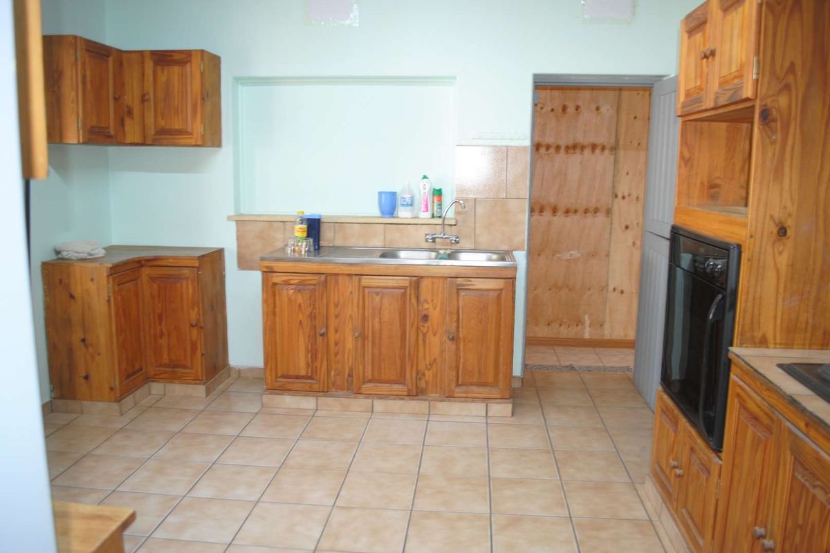 A dated kitchen featuring wooden cabinetry, tiled flooring, a stainless steel sink, and a built-in oven, with an adjacent unfinished utility area.