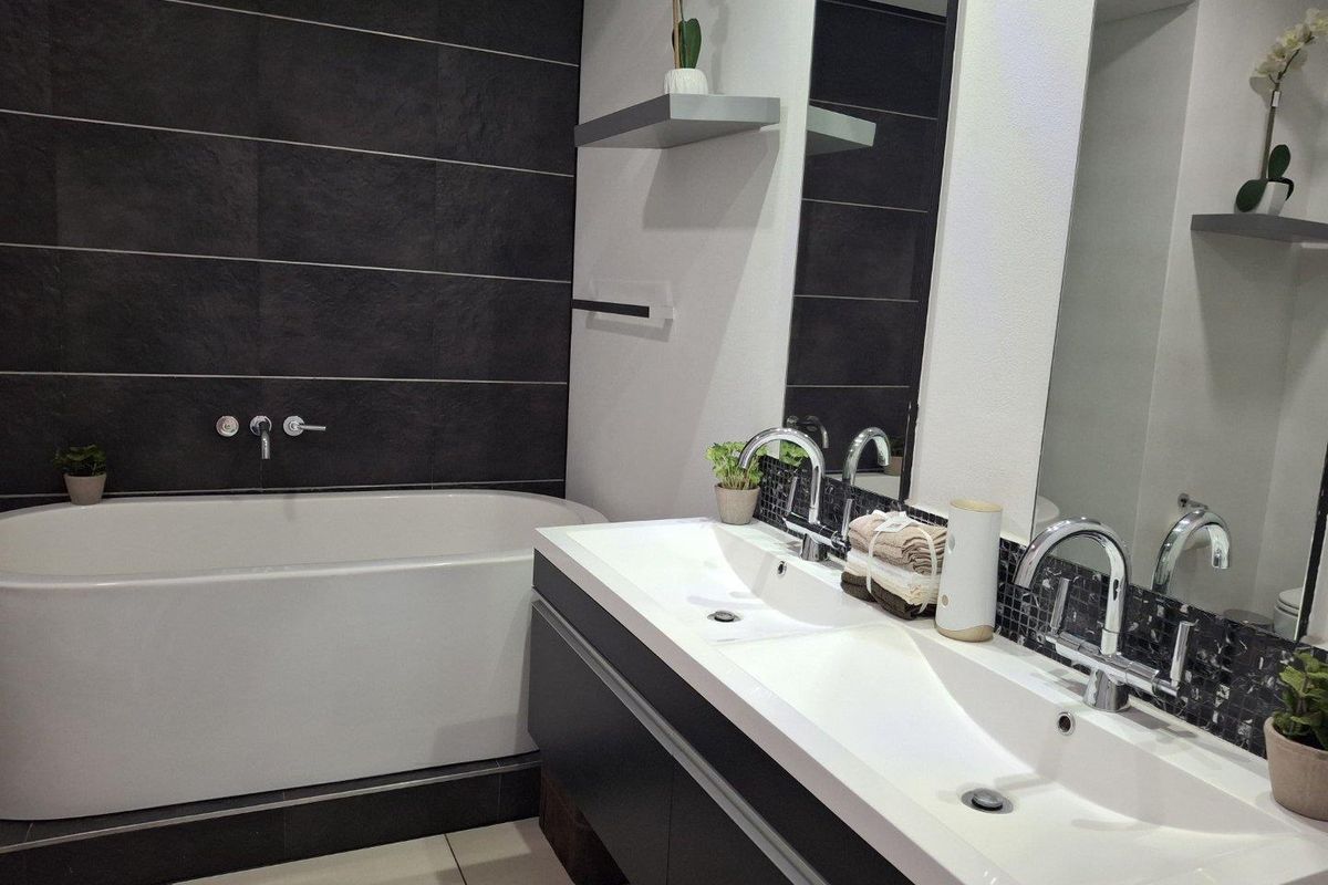 Modern bathroom featuring a sleek double vanity with integrated sinks, wall-mounted faucets, and a contemporary freestanding bathtub against a dark tiled feature wall.