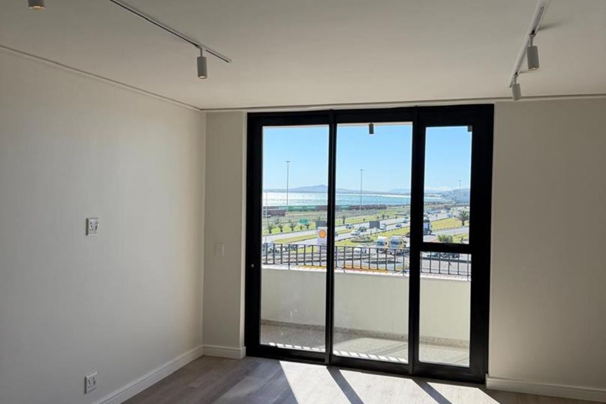 Bright and modern empty living area featuring laminate flooring, track lighting, and large sliding glass doors opening to a balcony with coastal views.