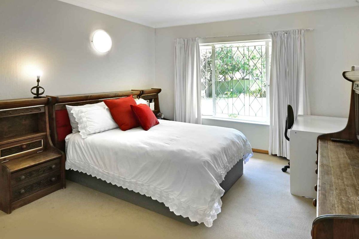 A well-lit bedroom featuring neutral carpeting, a large window with security bars, and a wall-mounted light fixture.