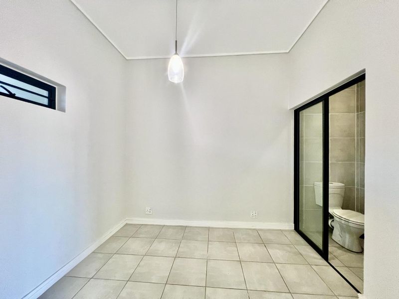 A bright, empty bedroom featuring neutral tiled flooring, a modern pendant light, and an en-suite bathroom accessed via a sleek black-framed sliding glass door.