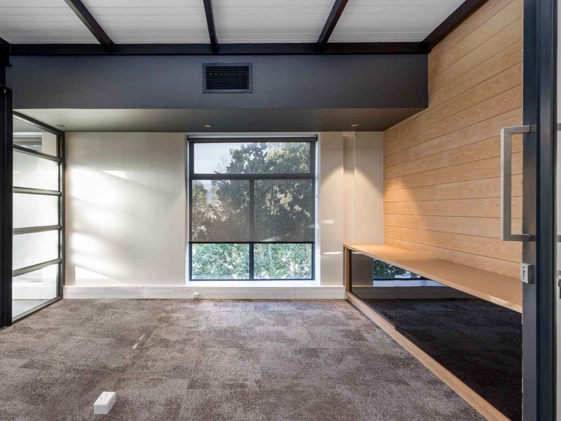A modern, empty office space featuring timber wall paneling, a built-in desk, exposed ceiling beams, and large windows with black metal frames.