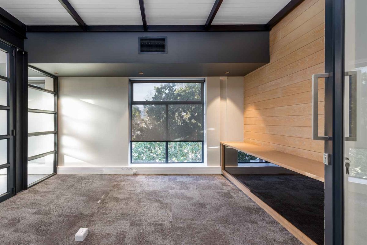 A modern, empty office space featuring timber wall paneling, a built-in desk, exposed ceiling beams, and large windows with black metal frames.