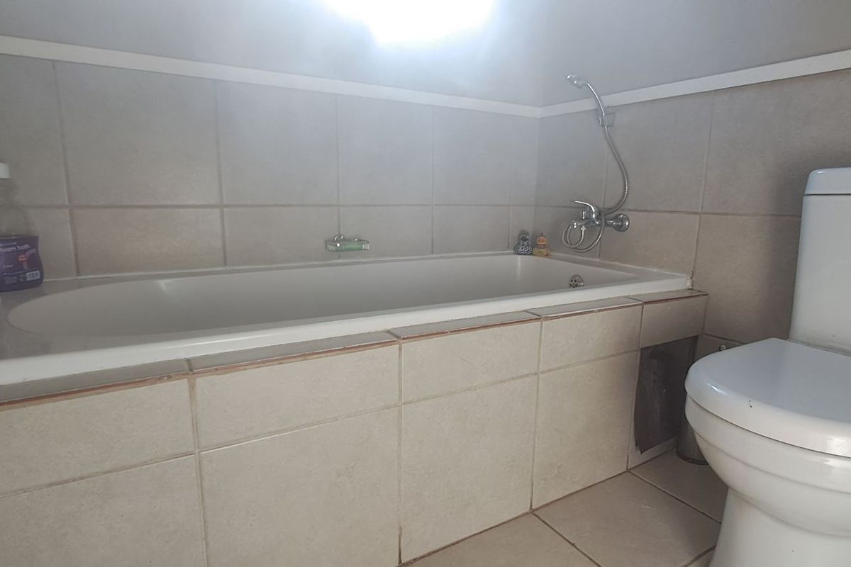 A functional bathroom featuring a built-in bathtub with a handheld shower mixer and a standard toilet, finished with neutral wall and floor tiling.