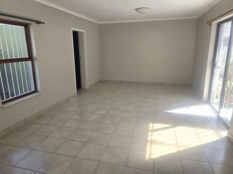 A bright, empty living room featuring neutral tiled flooring, a sliding glass door providing natural light, and security bars on the window.