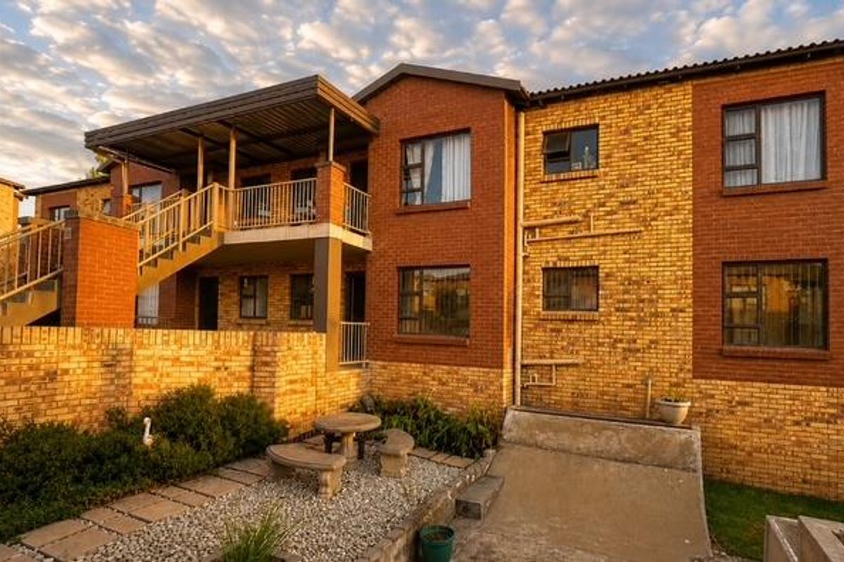 Rear view of a multi-story townhouse complex featuring brick facade, a raised balcony with metal staircase, and a paved patio area.