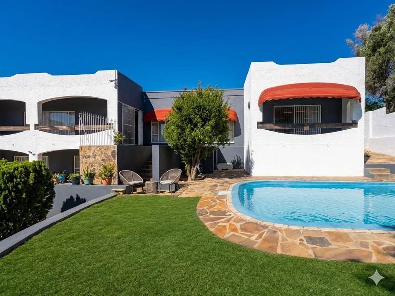 Rear exterior view featuring a swimming pool, stone paved surround, manicured lawn, and covered patio areas with red awnings.