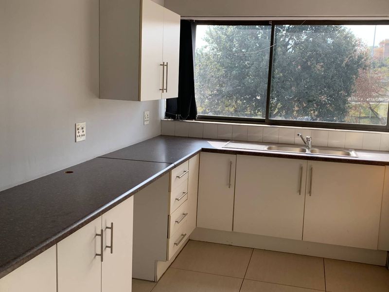 A functional office kitchenette featuring white cabinetry, dark laminate countertops, a stainless steel sink, and large windows providing natural light.