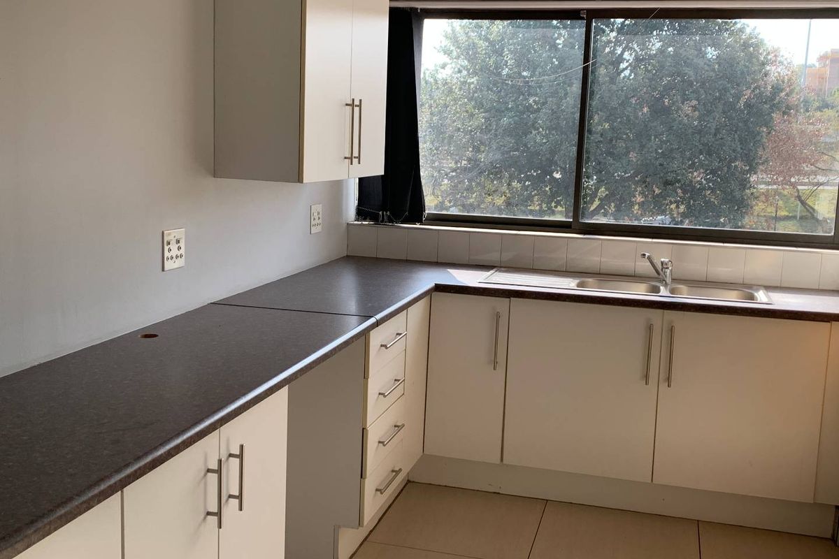 A functional office kitchenette featuring white cabinetry, dark laminate countertops, a stainless steel sink, and large windows providing natural light.