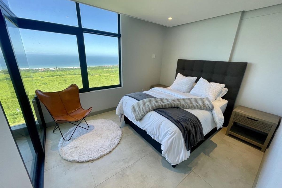 Modern bedroom featuring large floor-to-ceiling windows with expansive coastal and landscape views, neutral tiled flooring, and recessed ceiling lighting.
