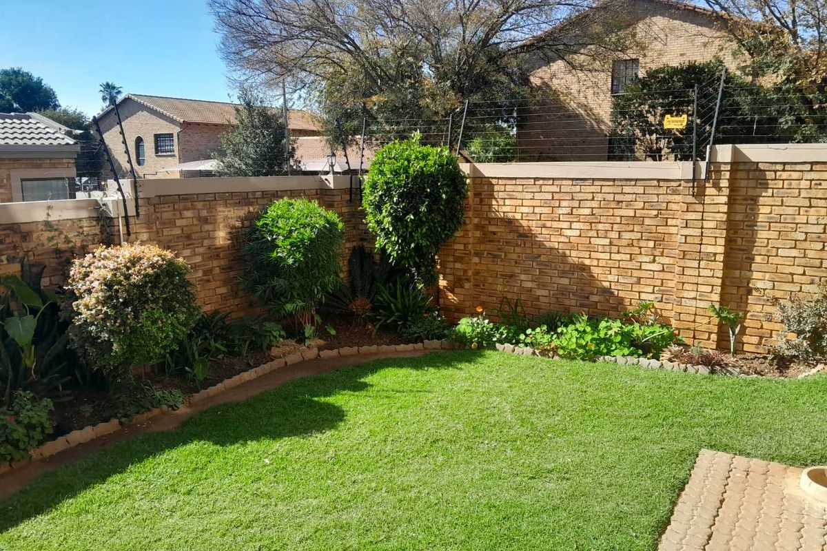 A private rear garden featuring a well-maintained lawn, landscaped garden beds, and a secure perimeter brick wall with electric fencing.