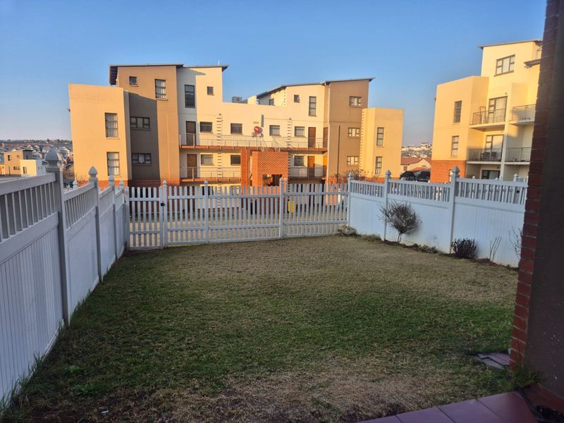 A private rear garden area featuring a well-maintained lawn enclosed by white vinyl fencing, set against a modern multi-story apartment complex.