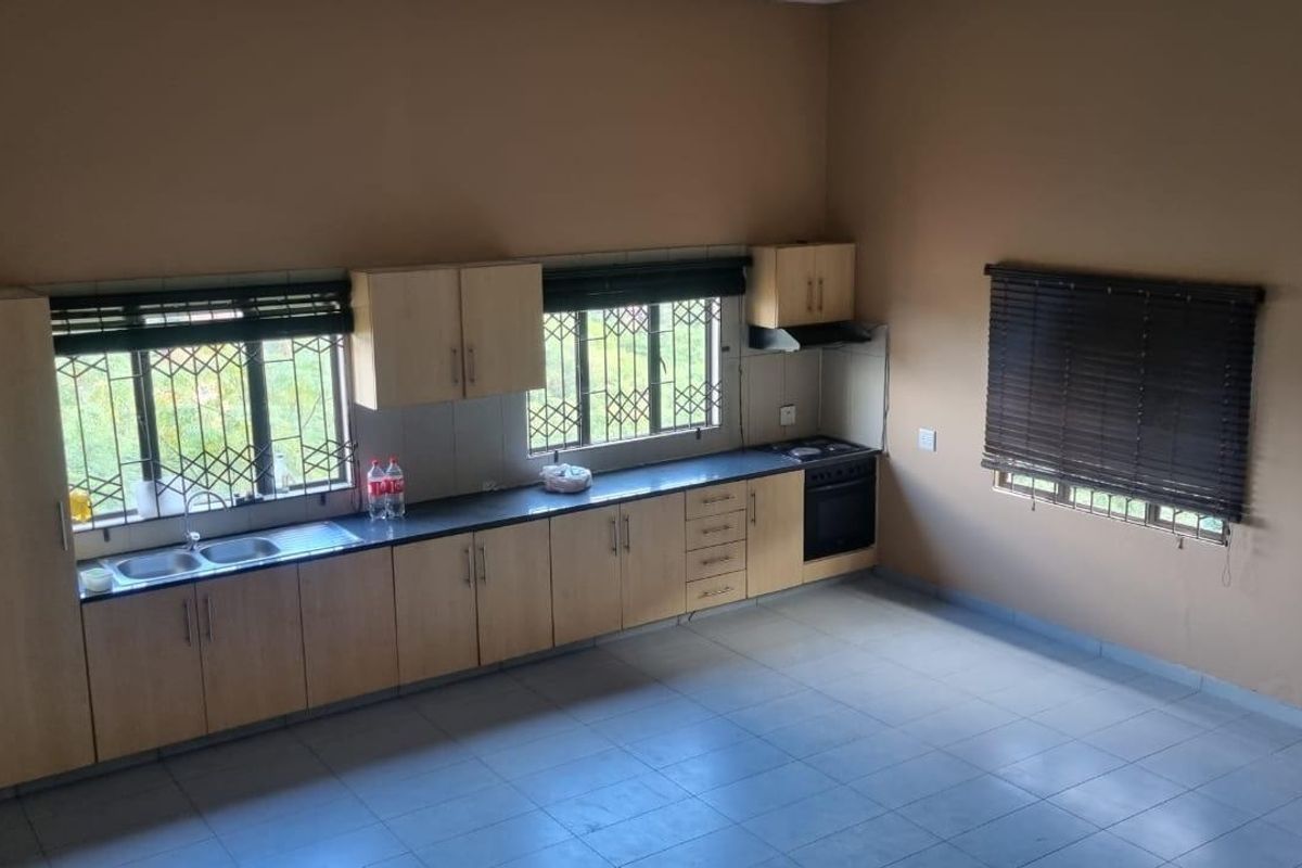 A spacious, empty kitchen featuring light wood cabinetry, dark countertops, built-in oven, extractor fan, and tiled flooring with large windows.