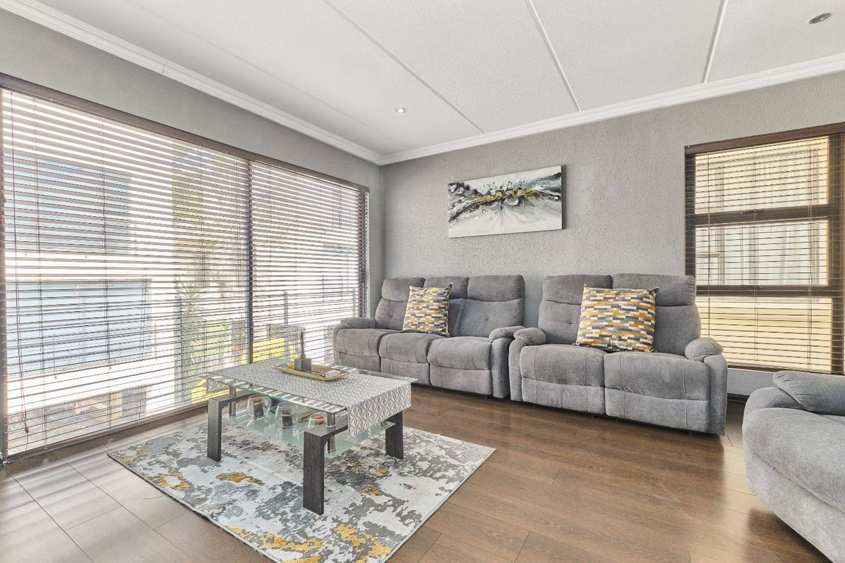 A well-lit living room featuring wood-look flooring, textured grey walls, and large windows fitted with horizontal blinds.