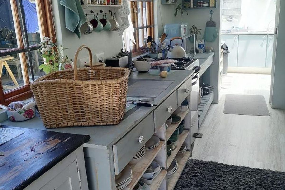 A rustic-style kitchen featuring open shelving, a gas hob, and a connected scullery area with light-toned cabinetry and timber-framed windows.