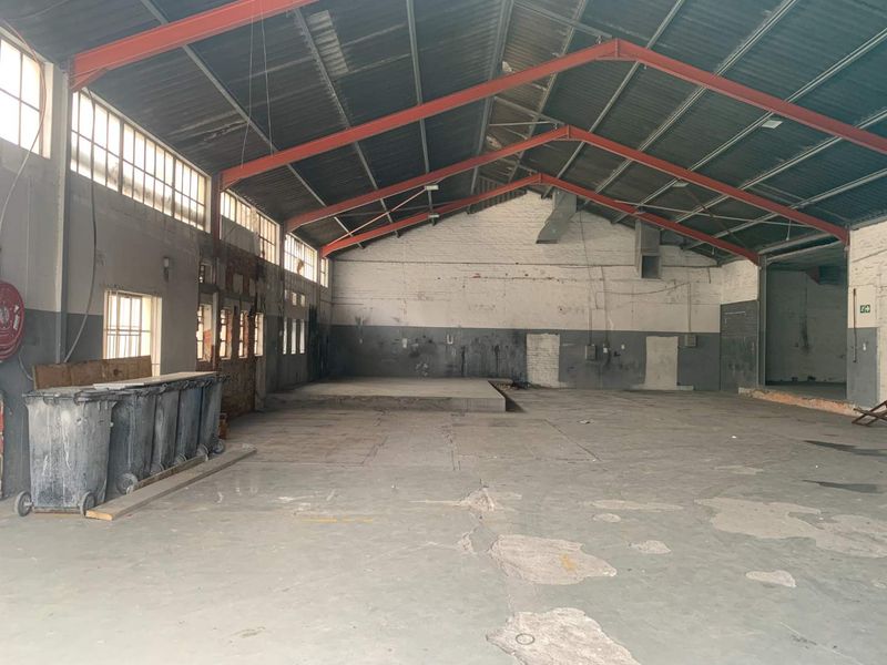 Spacious industrial warehouse unit featuring high ceilings with exposed steel trusses, concrete flooring, and large windows, suitable for various commercial or retail applications.