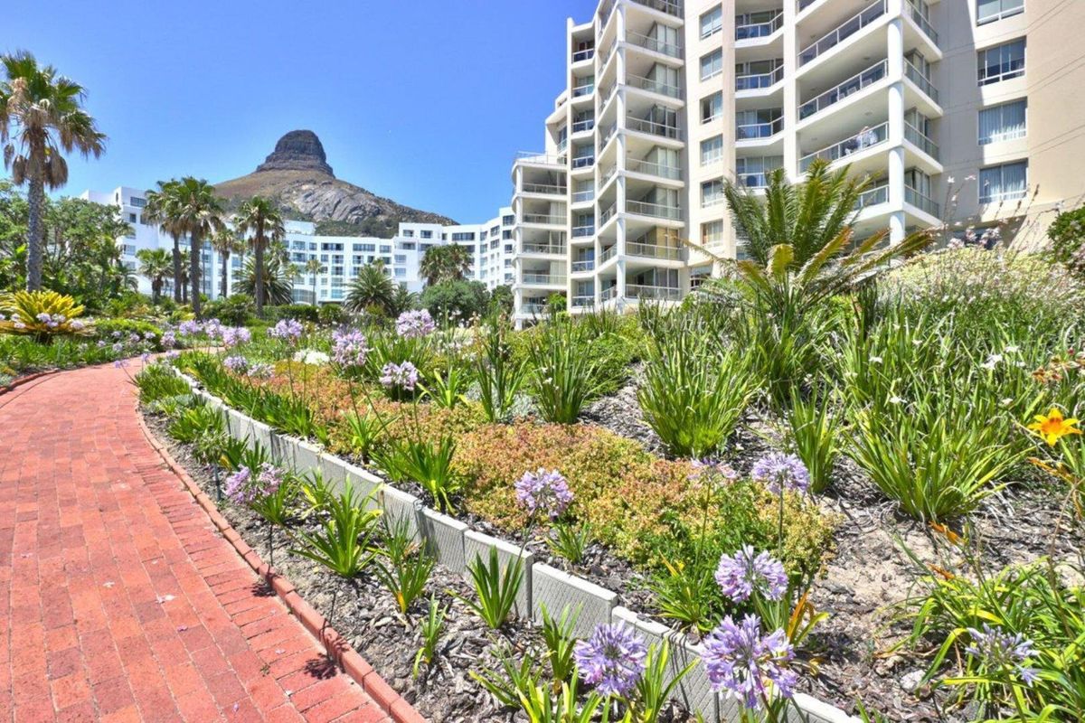 A well-maintained landscaped garden with a brick pathway leading towards a multi-story apartment complex with mountain views.