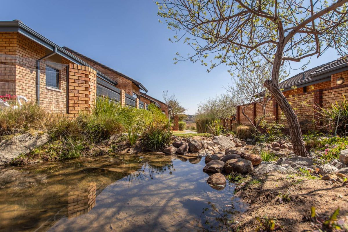 A well-maintained exterior rear view of a retirement village unit featuring face brick construction, a landscaped garden, and a natural rock water feature.