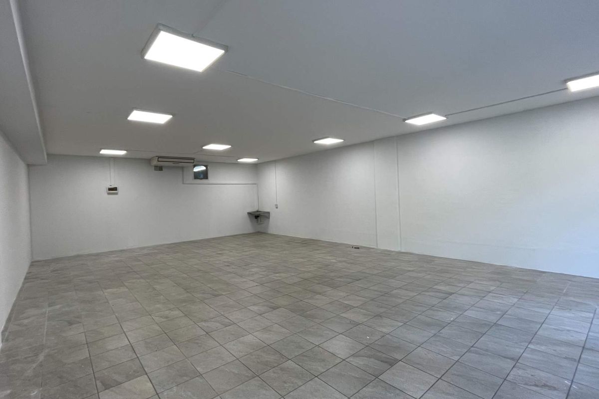 A spacious, empty open-plan retail area featuring neutral tiled flooring, recessed ceiling lighting, and a wall-mounted air conditioning unit.