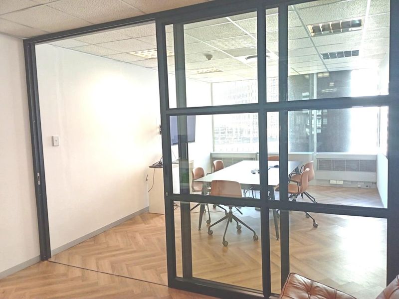 A modern office meeting room featuring a sleek black-framed glass partition, herringbone wood flooring, and large windows providing ample natural light.