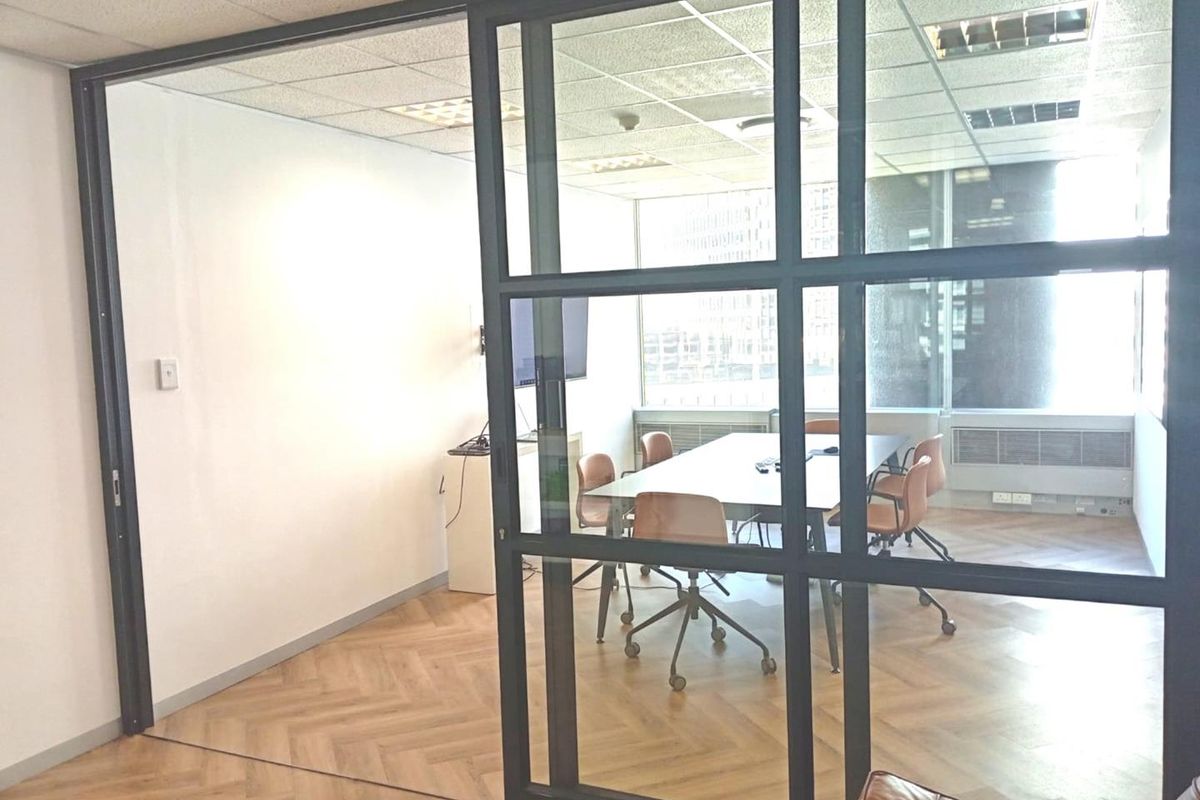 A modern office meeting room featuring a sleek black-framed glass partition, herringbone wood flooring, and large windows providing ample natural light.