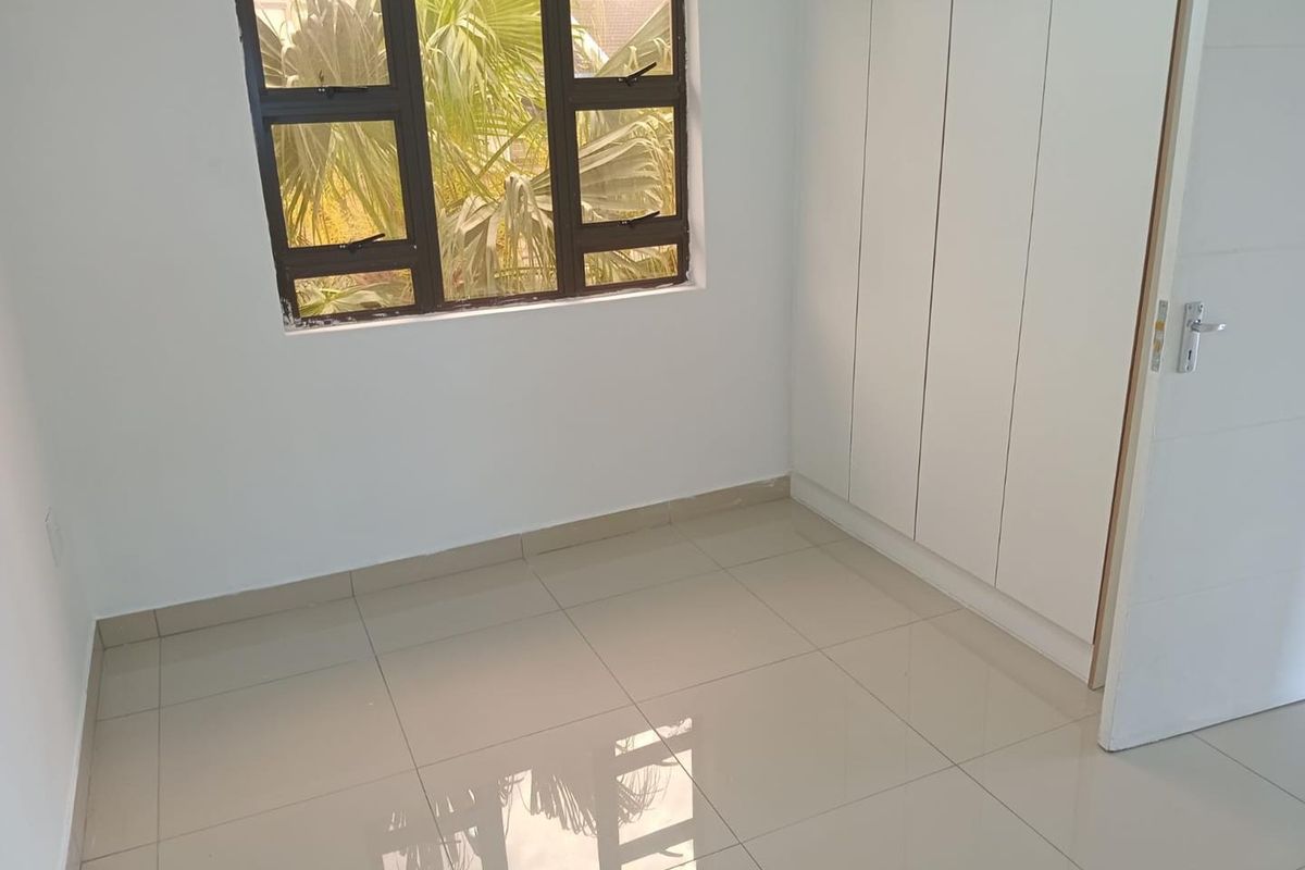 A clean, empty bedroom featuring neutral tiled flooring, built-in cupboards, and an aluminium-framed window providing natural light.