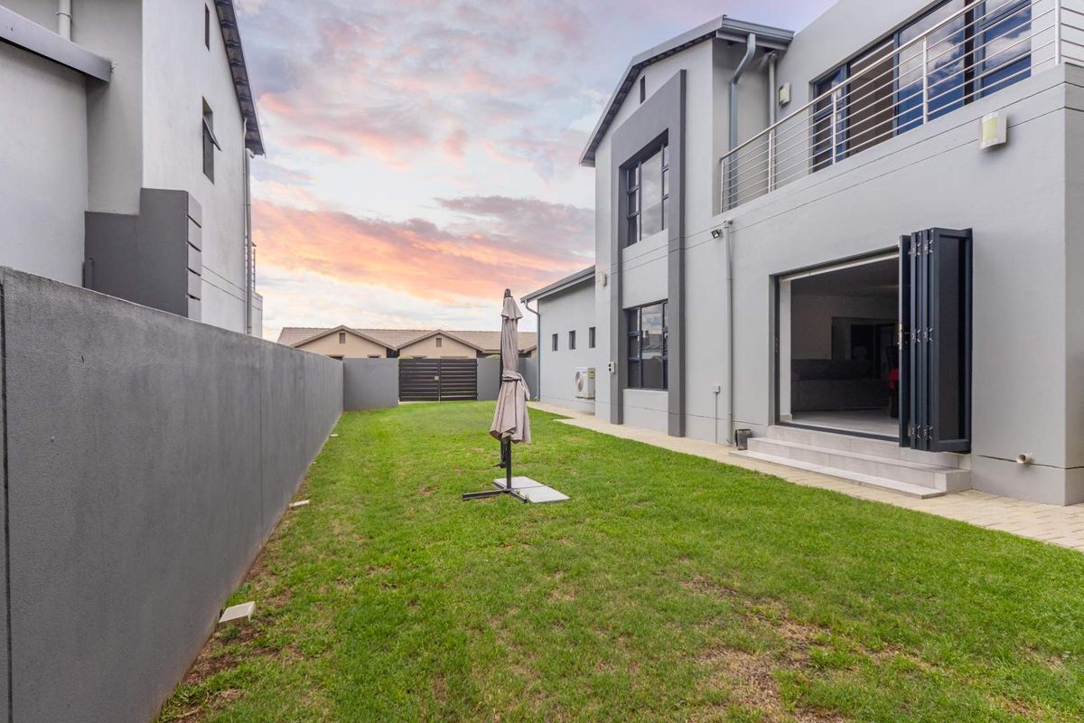 Rear view of a modern two-story house featuring a manicured lawn, boundary wall, paved patio area, and large bi-fold doors leading to the interior.